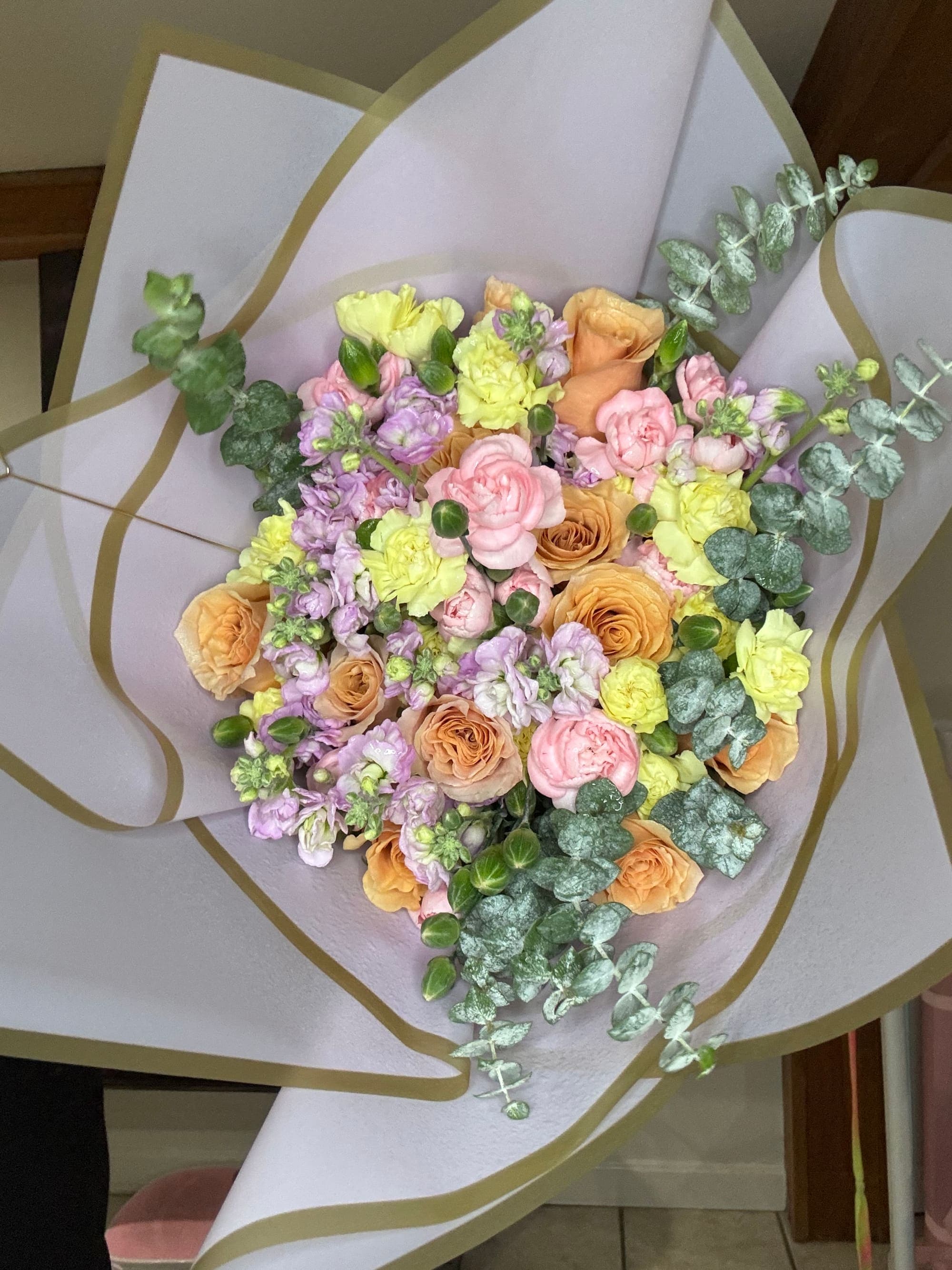 Handcrafted bouquet of peach roses, pink and yellow carnations, lavender stock and eucalyptus wrapped in lilac paper with a gold edge