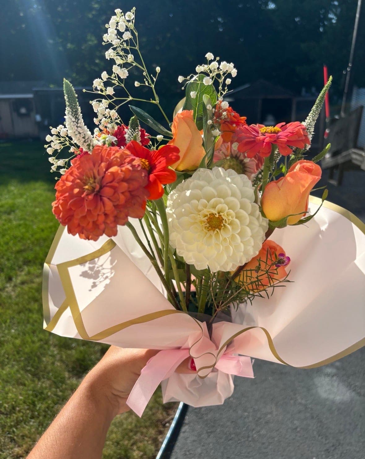 Hand-held bouquet of orange zinnias, a white dahlia, peach roses and baby’s breath wrapped in blush paper, glowing in afternoon sun