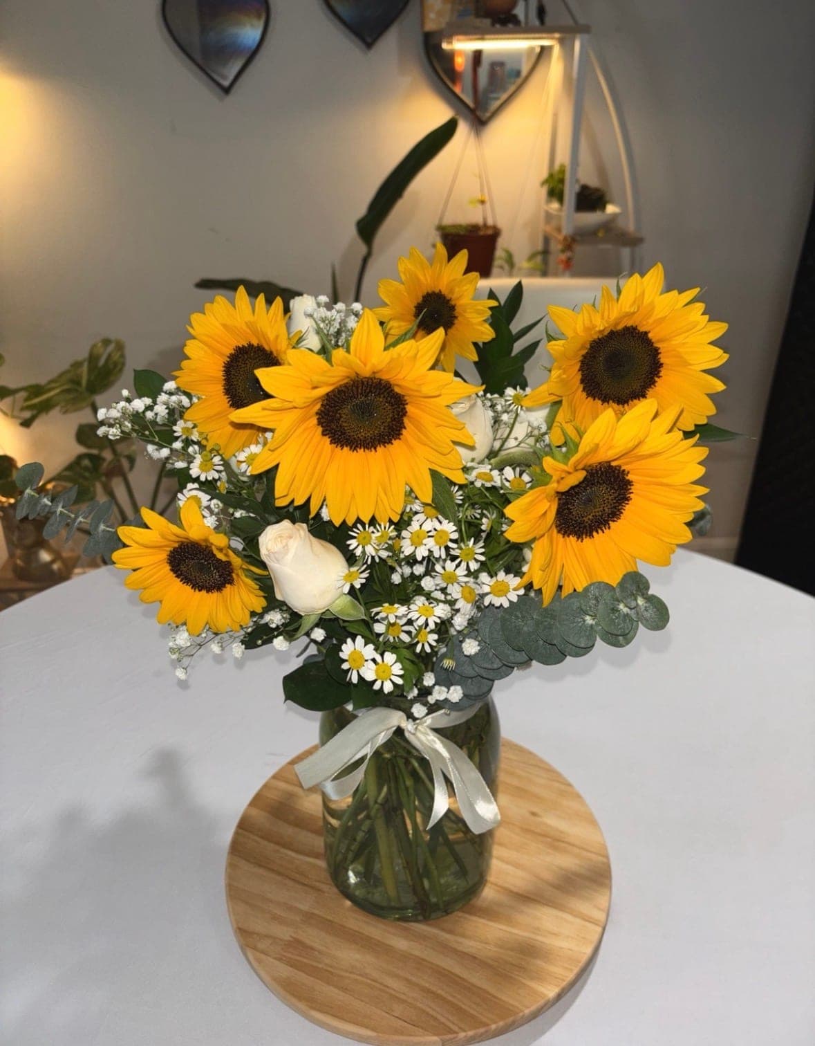 Vase arrangement of sunflowers, white roses, chamomile daisies and eucalyptus in a glass jar tied with a cream ribbon